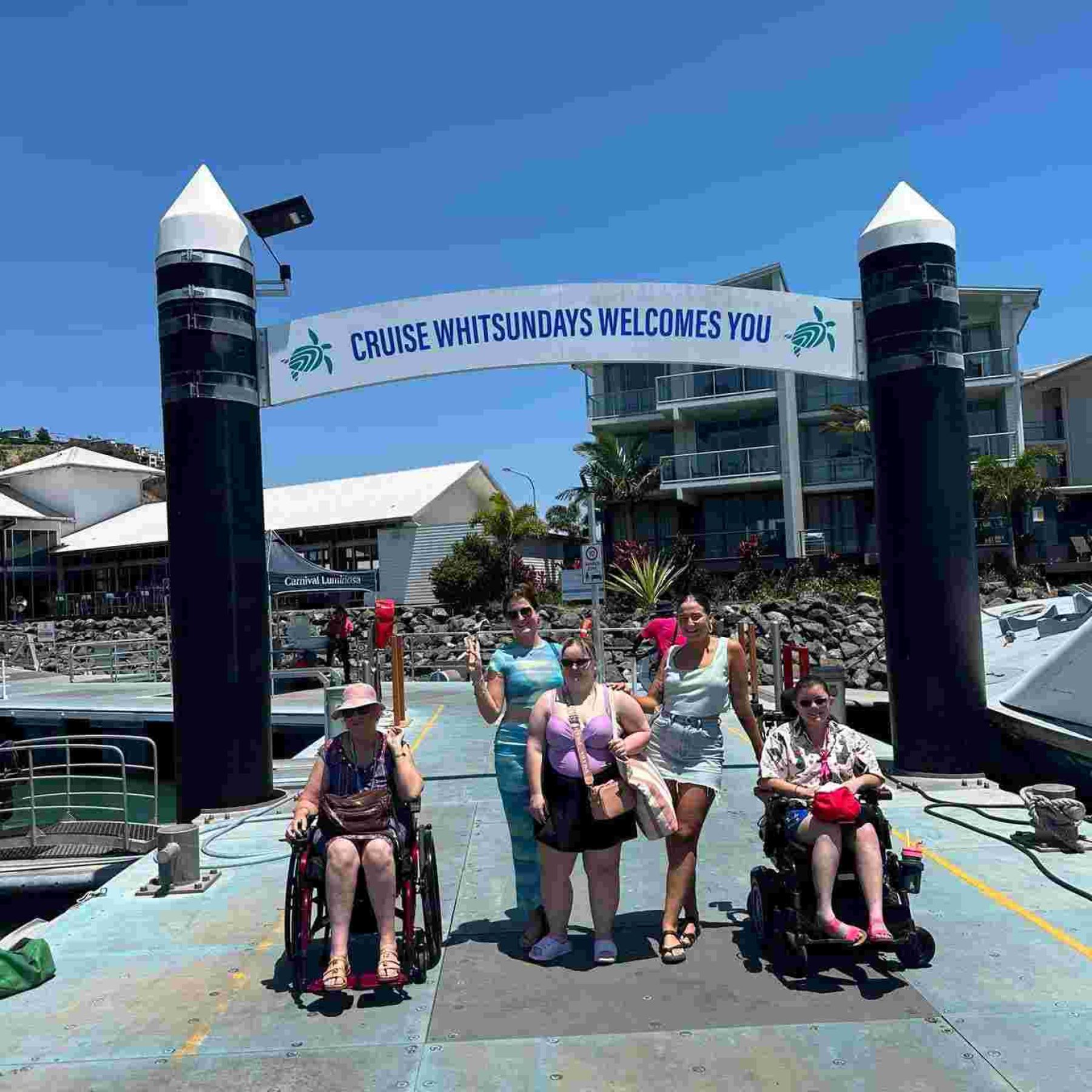 Group of people and two disabled people in their wheelchair smiling for a picture in front of cruise whitsundays welcomes you